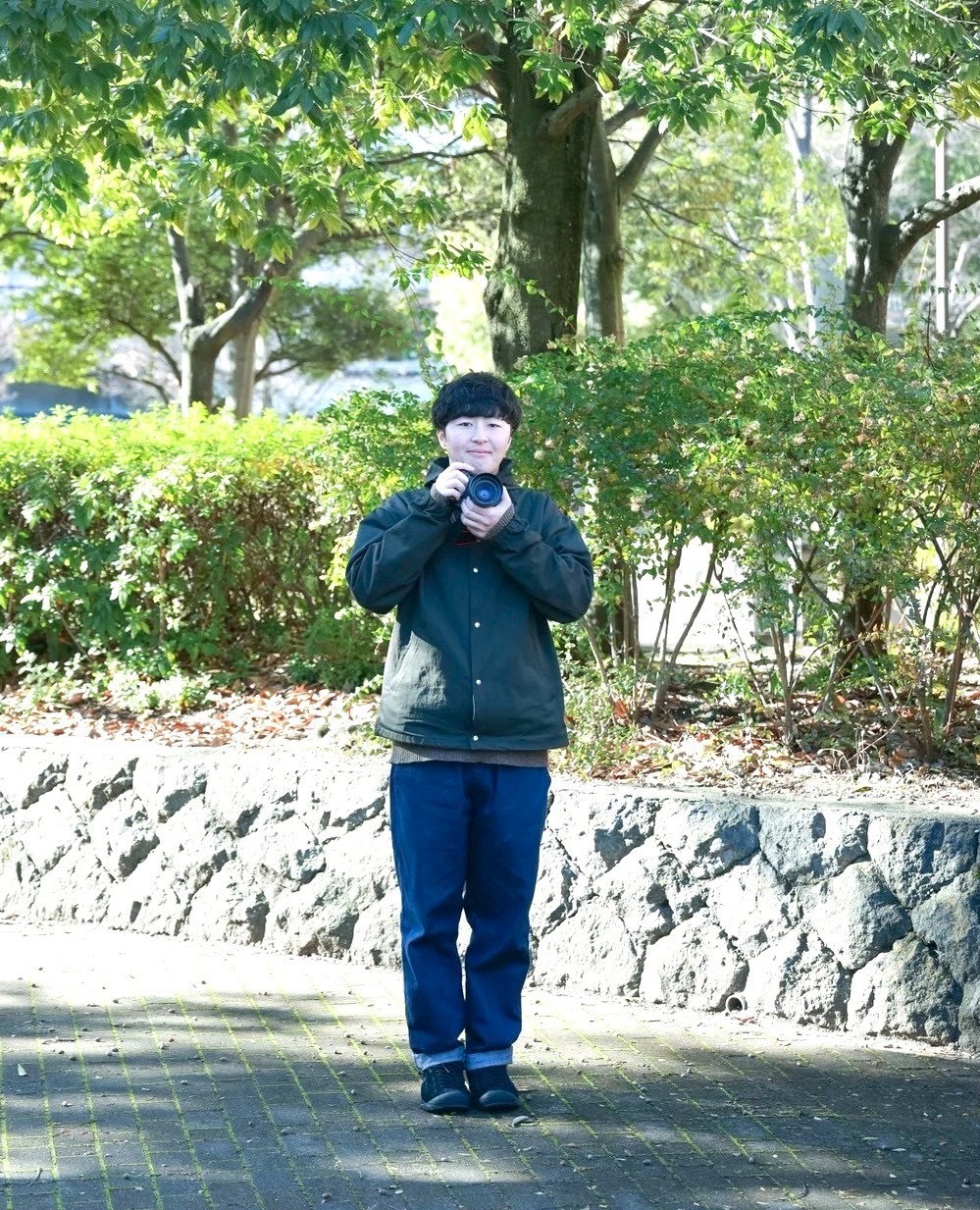 カメラマン湯本拓郎氏。中野市北公園にて木漏れ日の下でカメラを持っている姿。